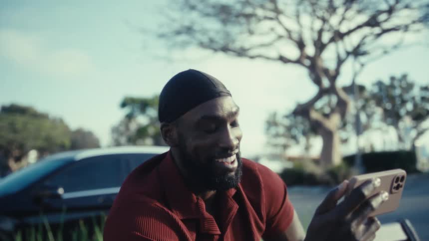 A black man wearing a cap and a polo shirt is seated outdoors on a bench looking up with a wide joyous smile while holding a mobile phone in his left hand and raising his right index finger to the sky against a backdrop of trees and a parked white