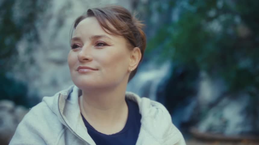 The face of an adult woman with a smile on her face in an outdoor setting looking ahead and behind her is a small waterfall