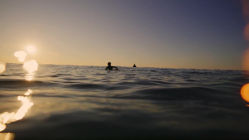 Two people sit on a surfboard riding across the ocean at sunset as gentle waves create rippling effects on the waters surface