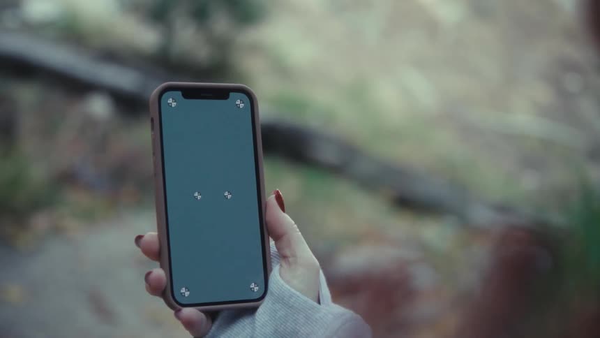 A woman holds a smartphone displaying a dark screen with white tracking markers in front of a blurred forest background