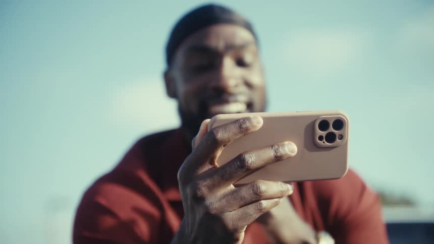 A young black man excitedly making a video call on his phone stands with a tree in the background at midday
