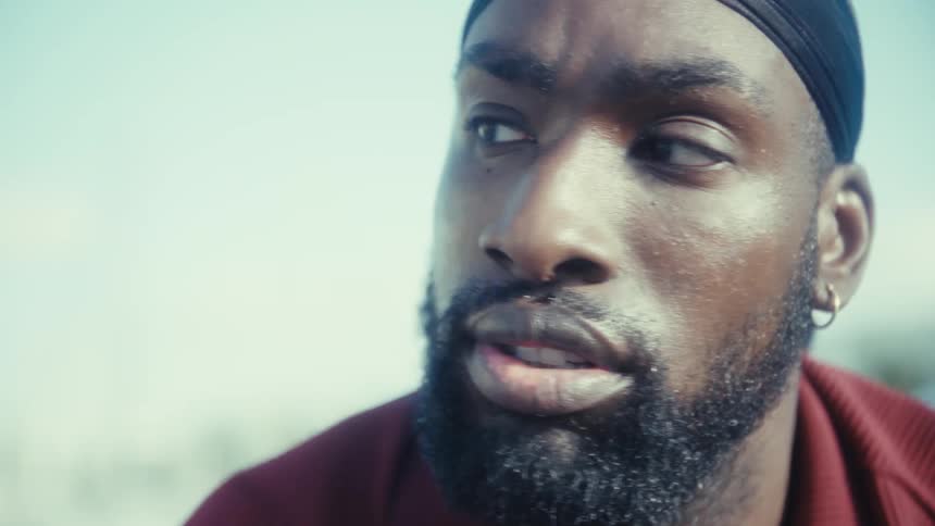 The video starts with a closeup of a man holding a smartphone in a tan case which he then lowers he is wearing a black durag and a red collared shirt his expression is serious as he looks up and gazes offcamera with sweat visible on his face