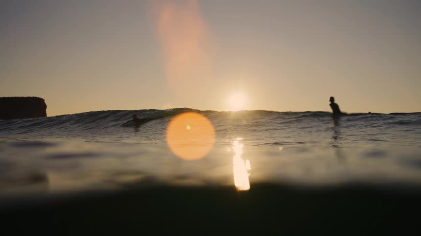 Two surfers paddle in the water at sunset with the ocean surface filling the foreground