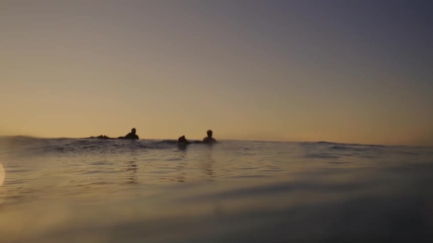 Two silhouetted surfers paddle in the ocean at sunset viewed from the waters surface