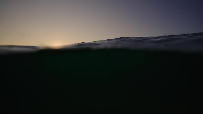 A halfsubmerged camera view shows the ocean at sunset capturing the dark water below the surface and the moving waves above with a bright sun setting near the horizon and a surfer in the distance