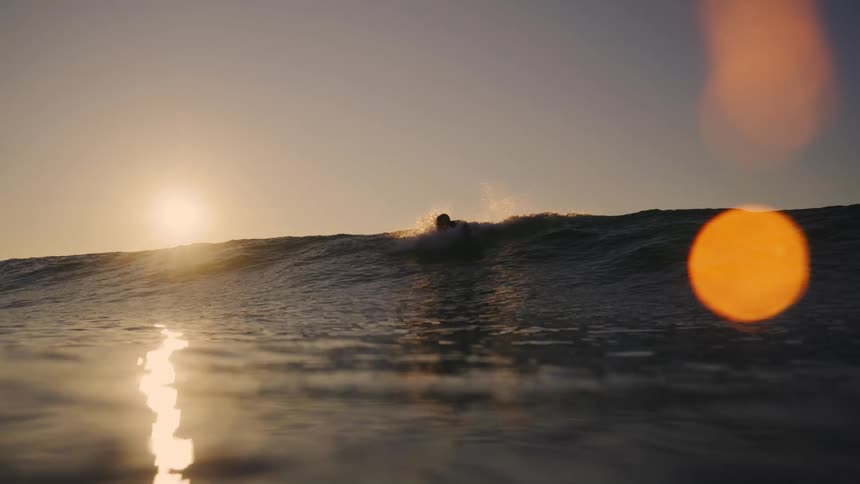 A man swims on a surfboard in the ocean at sunset heading toward the waves