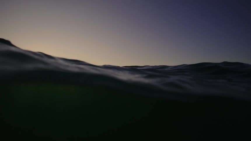 Two people sit on a surfboard in the distance out on the ocean at sunset as waves create rippling effects across the water