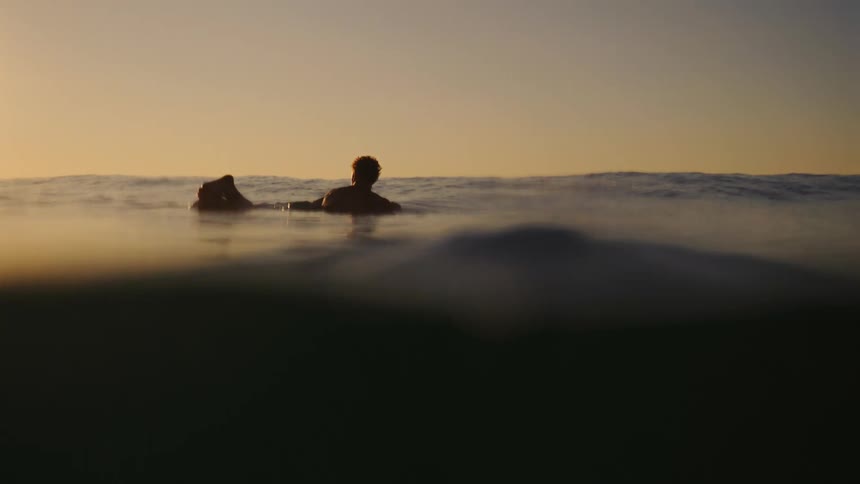 The video is a lowangle splitlevel shot capturing a surfer paddling out on a board in the ocean silhouetted against a bright warm sunset or sunrise skythe scene features strong lens flares and bokeh in the foreground water emphasizing the tranqu