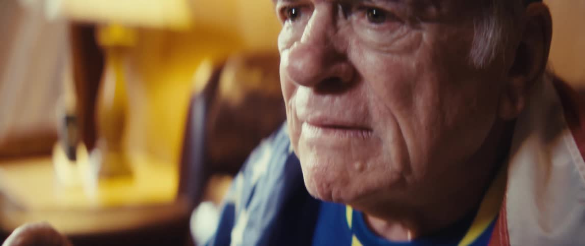 An elderly man excitedly watches sports on tv at home while wearing a flag