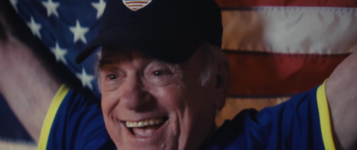 Elderly man wearing a baseball cap and a football jersey cheering and clapping with an american flag in a living room indoors during night time