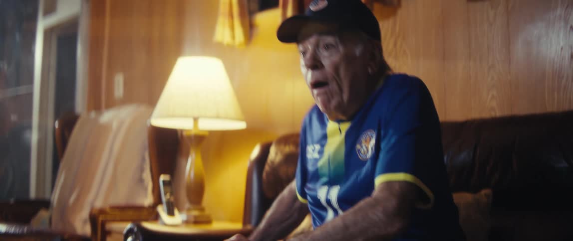 Elderly man wearing a baseball cap and football jersey sitting on a couch laughing and cheering while holding an american flag indoors during day time