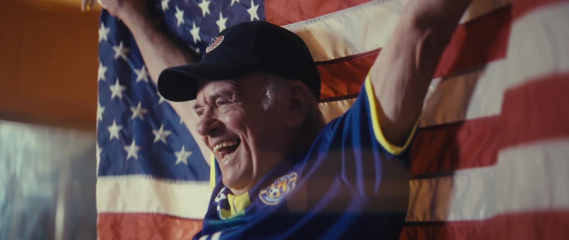 Elderly man wearing a baseball cap and a football jersey cheering and dancing while holding an american flag in a living room indoors during night time