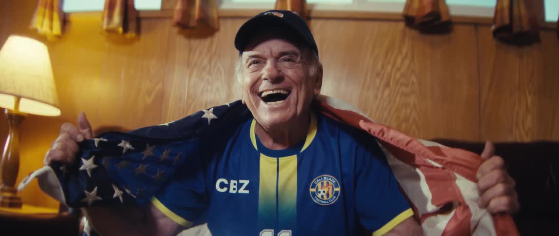 An elderly man sits excitedly on a couch in his living room a projector in front of him holding an american flag while wearing a jersey and a hat as he watches a sports game