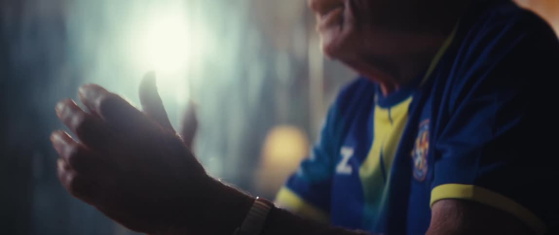 An elderly man wearing a hat and a sports jersey cheers enthusiastically for his team while sitting in his living room watching the game