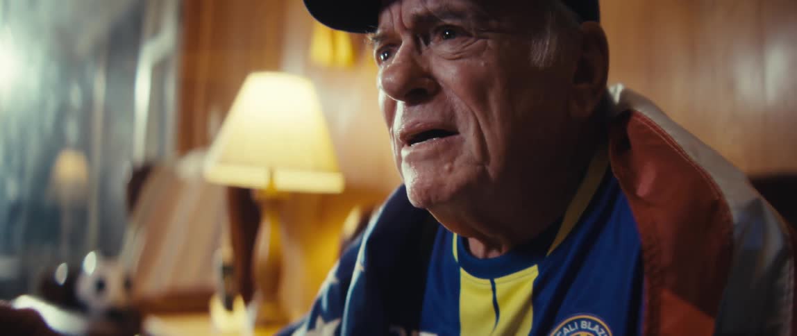 An elderly man wearing a jersey and a hat wraps a us flag around himself as he sits on a couch in his living room watching a sporting event with excitement