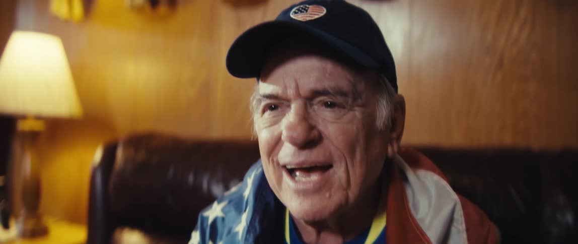 An enthusiastic elderly man draped in an american flag claps and speaks animatedly while wearing a cap with a heartshaped flag patch