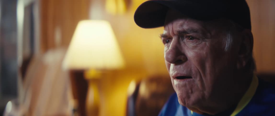 Elderly person in dark cap and blue shirt with yellow lanyard in warmly lit cozy indoor setting