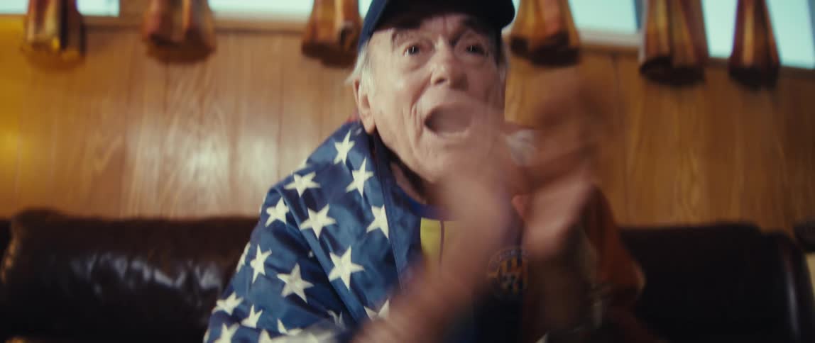 Focus shifts from a movie projector to an excited elderly man draped in an american flag cheering wildly