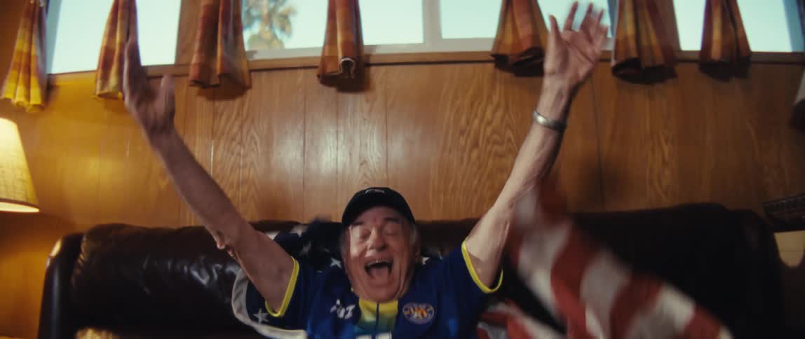 An elderly man wearing a jersey and draped in an american flag leaps off his couch in excitement while watching a sports game projected in his living room