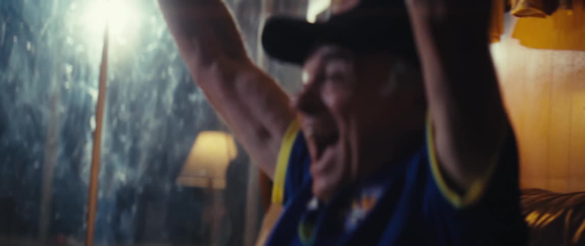 An elderly man wearing a hat and a sports jersey sits on his couch in the living room screaming with excitement as he watches the game
