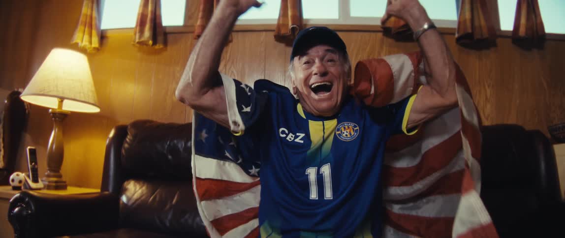 An elderly man dressed in a jersey wraps a us flag around himself while sitting on a couch in his living room watching a sporting event with excitement