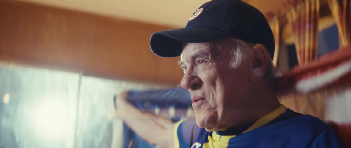 An elderly man dressed in a sports jersey sits in his living room wrapped in an american flag his face lit with excitement as he watches a game