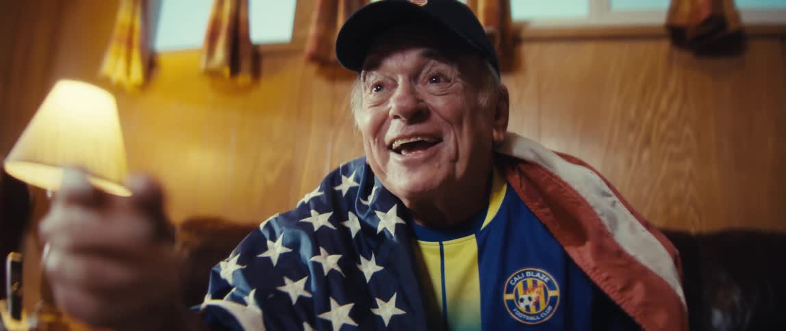 Elderly man wrapped in an american flag wearing a baseball cap and sports shirt cheering and clapping in front of a projector indoors