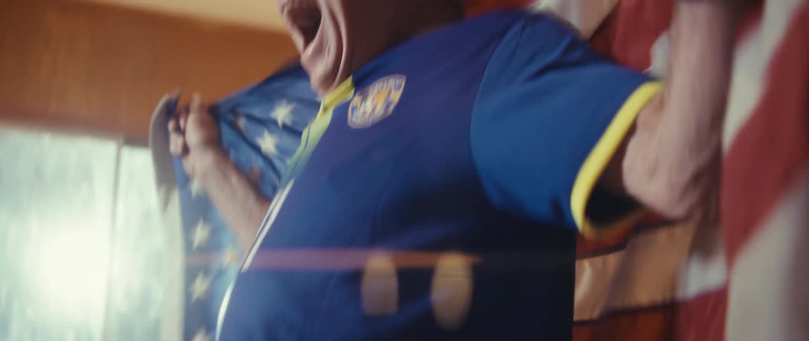An elderly man wearing a sports jersey and a hat stands up from his couch in excitement draped in an american flag as he celebrates the game