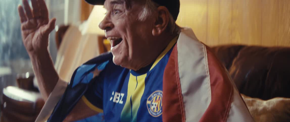 Elderly man dressed in a football jersey with a an american flag wrapped around his body cheering and clapping sitting in his sofa indoors during night time