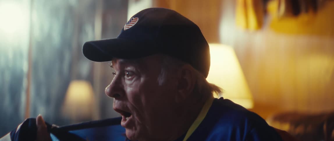 This video shows an older man dressed in a sports jersey and yellow socks sitting on a leather couch while covered with an american flag he transitions from a moment of intense concentration to cheering loudly and celebrating suggesting he is passiona