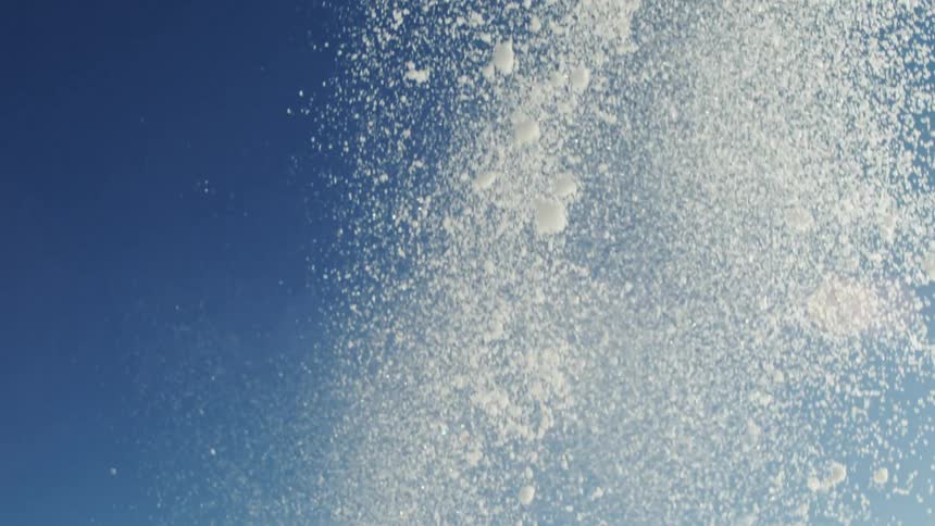 The video shows a dense shower of white particles which appear to be snow or ice crystals falling against a clear bright blue sky the sunlight catches the falling particles and a distinct lens flare is visible in the background suggesting a sunny wi