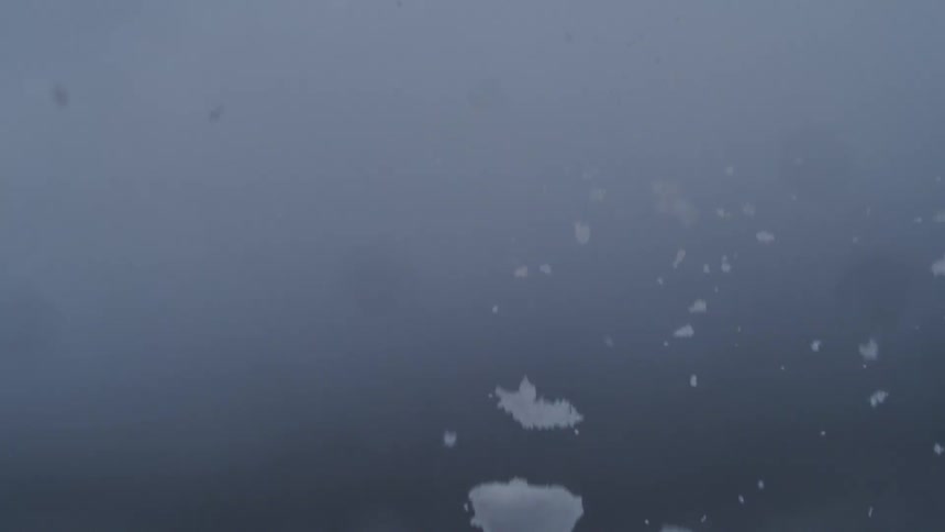 The video features a static shot against a hazy uniform bluegray background which suggests an overcast day or heavy fogmist white irregular particles likely large chunky snowflakes or soft hailgraupel fall rapidly and continuously through the fr