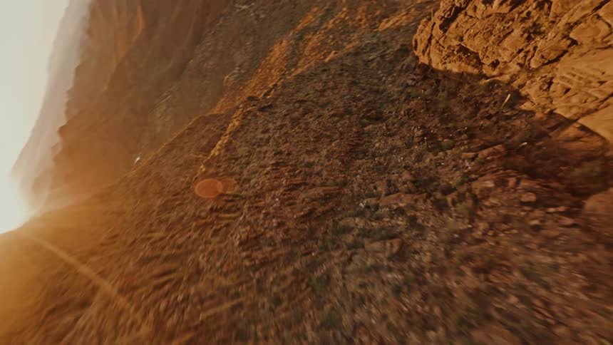 This is a highspeed dynamic firstperson view shot moving rapidly down and along the side of a steep rocky mountain or canyon face the video captures the texture of the dry brown terrain with the bright sunlight flaring over the ridge in the backgro