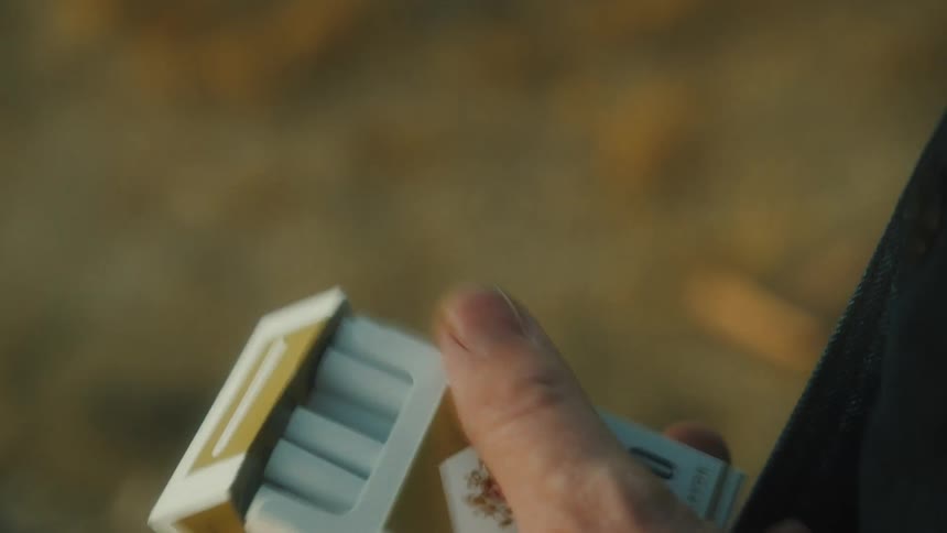The video is a closeup shot focusing on a persons hands opening a packet of cigarettes specifically tearing the foil or paper seal at the top the background is blurred and appears to be an outdoor setting with earthy or muted colors