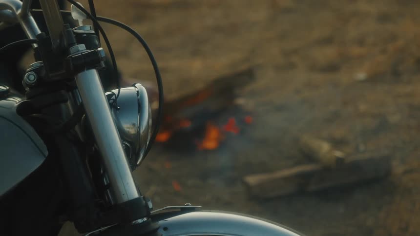 The video is a closeup on the front section of a motorcycle showing the headlight handlebars and front fender in the blurred background a small campfire is burning on the ground suggesting a roadside stop or campsite at dusk or night