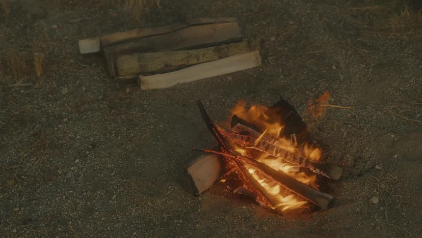 This overhead shot captures a small contained campfire burning brightly on dry dusty ground with the flames illuminating the stacked firewood several unburnt wooden planks are resting nearby ready to be added to the fire