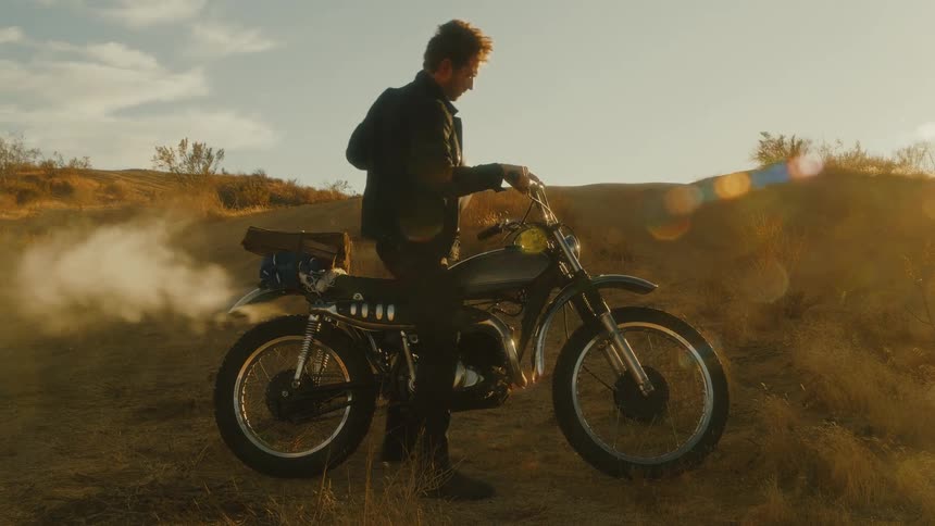 A man kick starts a vintage motorcycle creating a puff of white smoke and then picks up his helmet in the golden sunlight
