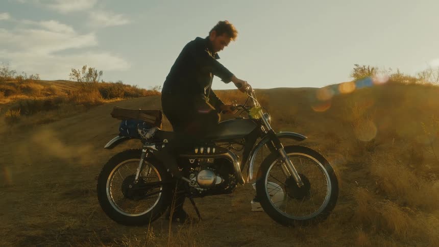 A man kick starts a vintage motorcycle causing it to puff white smoke in a golden sunlit landscape