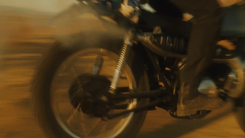 A close view of a rider accelerating a motorcycle up a steep dry hill kicking up dust against a backdrop of desert mountains and dramatic clouds