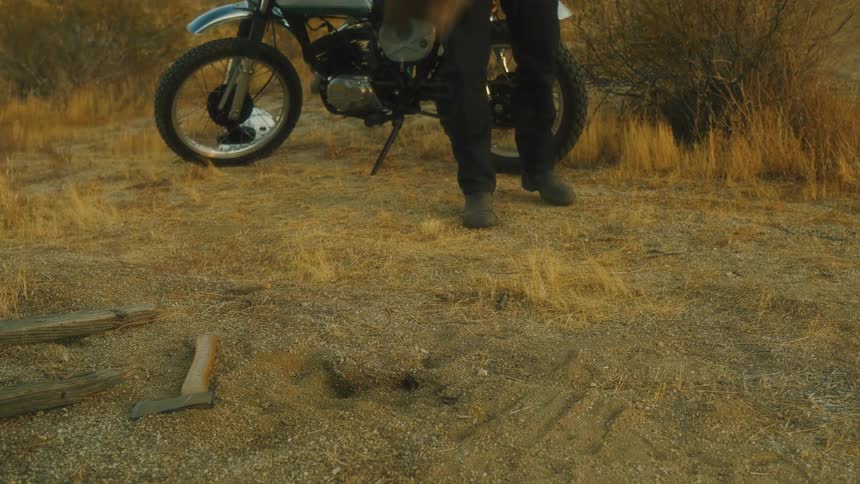 A person from the legs down is standing next to a parked motorcycle throwing logs to the ground near dry trees and grass