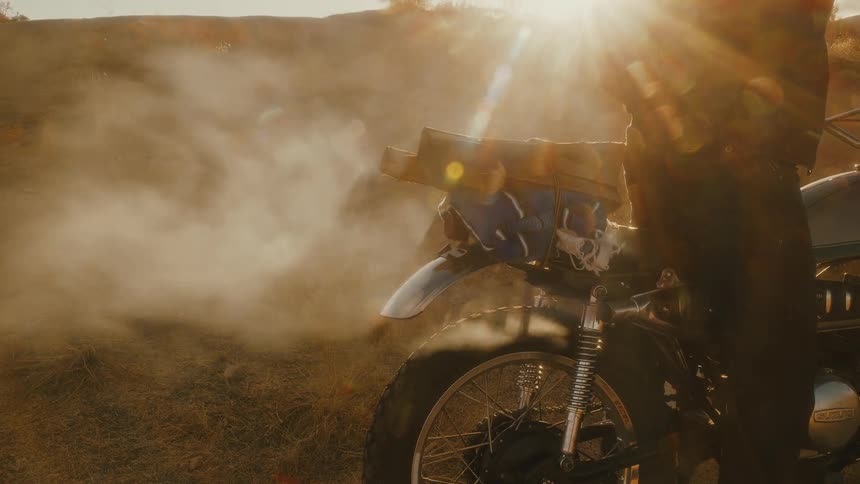 The video is a closeup shot focused on the front section of a parked motorcycle likely a vintage or scrambler style in a dusty sunlit outdoor setting smoke or dust billows dramatically around the bikes front tire and engine area with a strong lens
