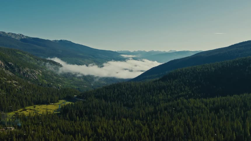 This video provides a breathtaking aerial view of a deep vshaped mountain valley densely covered in evergreen pine forests wisps of lowhanging mist or cloud drift over a small bright green clearing or meadow near the bottom of the valley with rocky