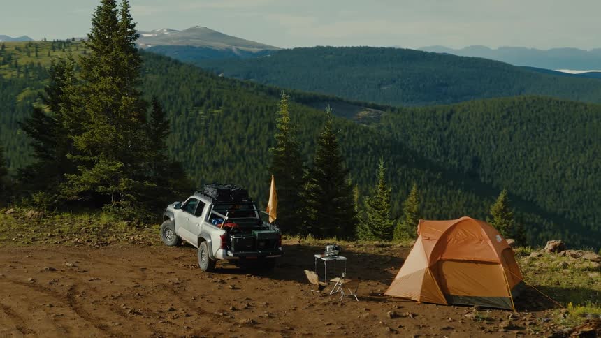 Aerial view of a truck and orange tent set up for camping above a vast forested mountain valley