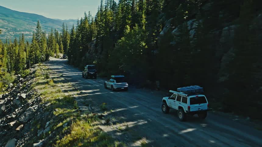 Three vehicles travel mountain road between rocky slope and pine forest