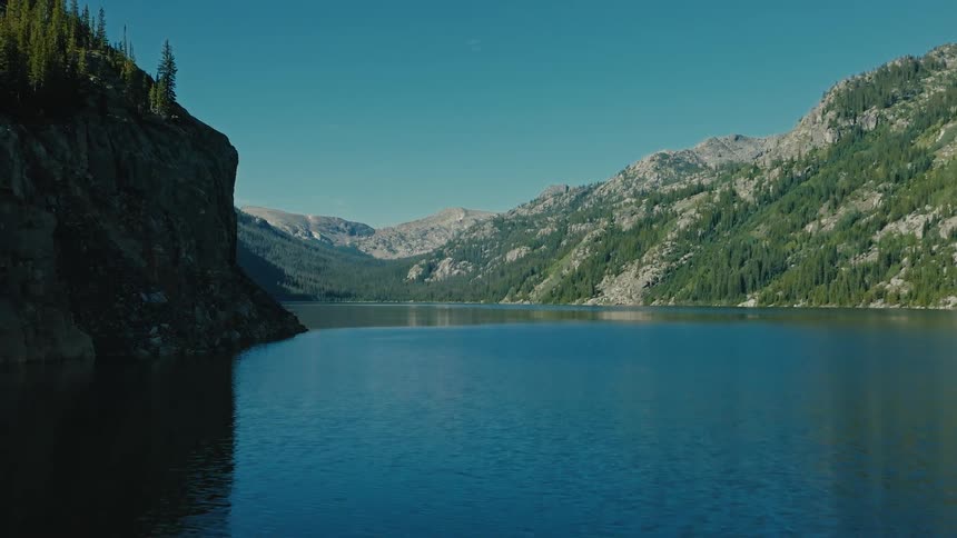 Blue water of a mountain lake is framed by a sheer dark cliff and rugged peaks covered in dense evergreen forest under a bright blue sky