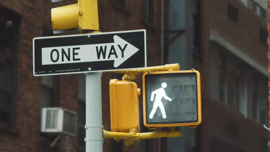The video features a closeup shot of a traffic pole displaying a black and white one way sign with an arrow pointing right attached beneath it is a yellow traffic signal box with a brightly lit pedestrian crossing signal white walking man indicating