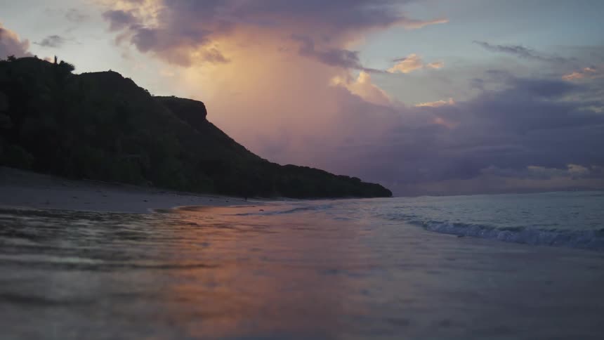 Soft waves lap against the shore at twilight as a dramatic cliff rises in the distance against a sky of purple clouds and fading orange light