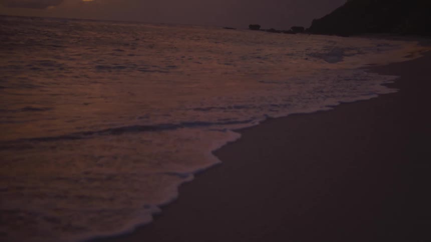 Gentle waves move across sand under orange evening sky before rocky coastline
