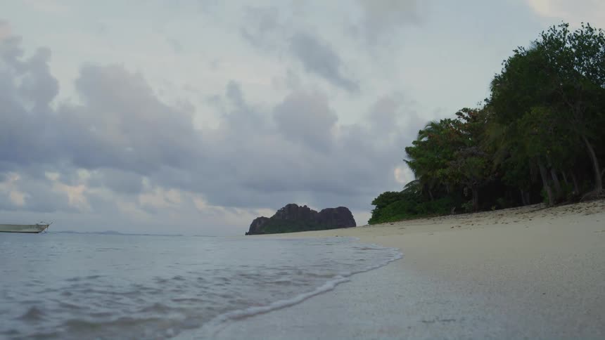 Waves crash against the beach on a cloudy day with trees lining the shoreline in the distance