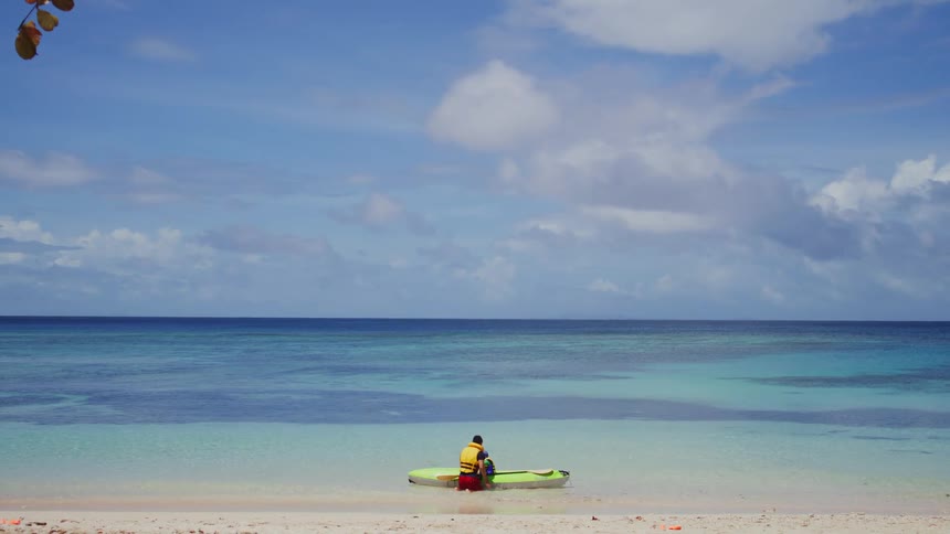 A person wearing a yellow life jacket sits on a green kayak at the edge of a white sand beach facing the calm turquoise ocean under a vast blue sky